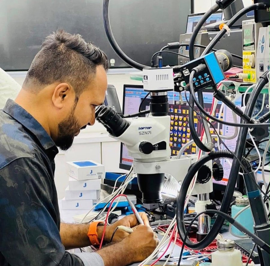 Moin Saiyad at the E11EVEN bench, working through a trinocular microscope with a 4K camera, bench power supplies and schematic screens around him